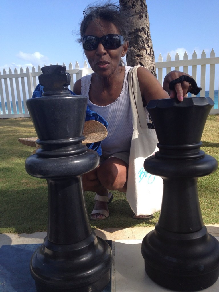 My mother, Carolyn, squatting next to life-size black king and black queen chess pieces on a Barbados beach.
